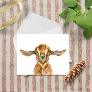 May include: A rectangular greeting card featuring a watercolor illustration of a brown baby goat with large, wide-set ears and a soft, textured coat. The goat is centered on a white background, looking directly forward with a gentle expression.