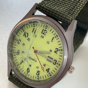 May include: A military-style wristwatch with a cracked, yellow-green face and a dark grey metal case. The watch has black numerals and hands, with a red second hand. It features a green nylon strap.