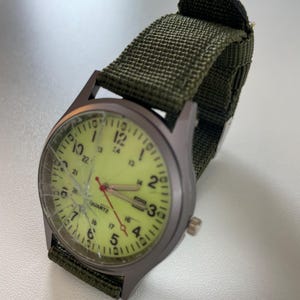 May include: A wristwatch with a green canvas band and a gray metal case. The watch face is a pale yellow color with black numerals and hands. The watch face is cracked. The word "QUARTZ" is printed on the watch face.