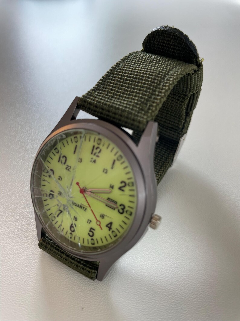 May include: A wristwatch with a green canvas band and a gray metal case. The watch face is a pale yellow color with black numerals and hands. The watch face is cracked. The word "QUARTZ" is printed on the watch face.