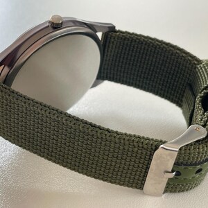 May include: Close-up of a wristwatch with a dark gray case and a green nylon strap. The watch has a silver buckle and a white watch face. The strap is approximately 0.75 inches wide.