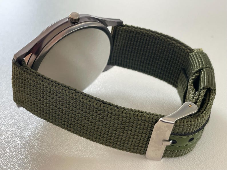 May include: Close-up of a wristwatch with a dark gray case and a green nylon strap. The watch has a silver buckle and a white watch face. The strap is approximately 0.75 inches wide.