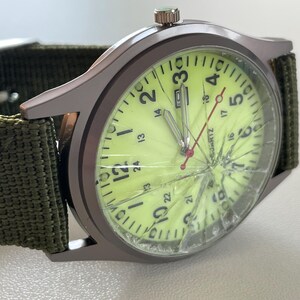 May include: A wristwatch with a cracked face and a green canvas band. The watch has a light green face with black numerals and hands. The watch case is a dark gray color.
