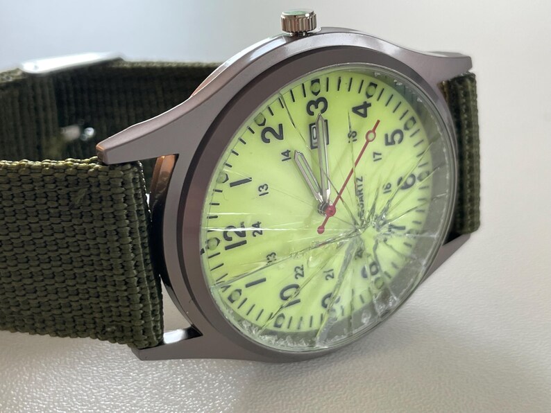 May include: A wristwatch with a cracked face and a green canvas band. The watch has a light green face with black numerals and hands. The watch case is a dark gray color.