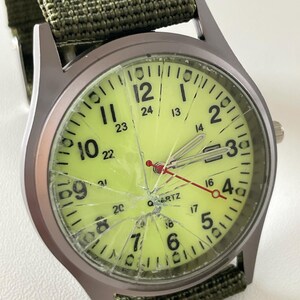 May include: A wristwatch with a cracked, light green face and a green nylon band. The watch has black numerals and hands, with a red second hand. The watch case is silver-toned.