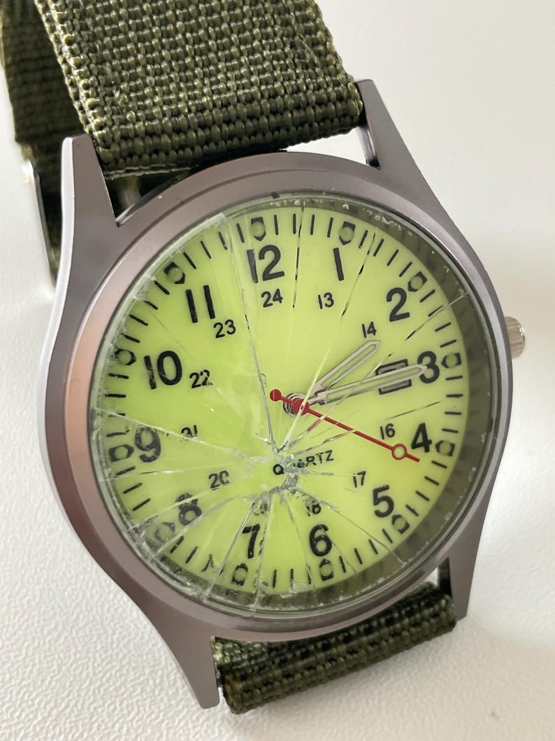 May include: A wristwatch with a cracked, light green face and a green nylon band. The watch has black numerals and hands, with a red second hand. The watch case is silver-toned.