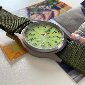 May include: A silver-toned wristwatch with a cracked, light green face and black numerals. The watch has a green nylon strap. The watch is resting on a photograph.