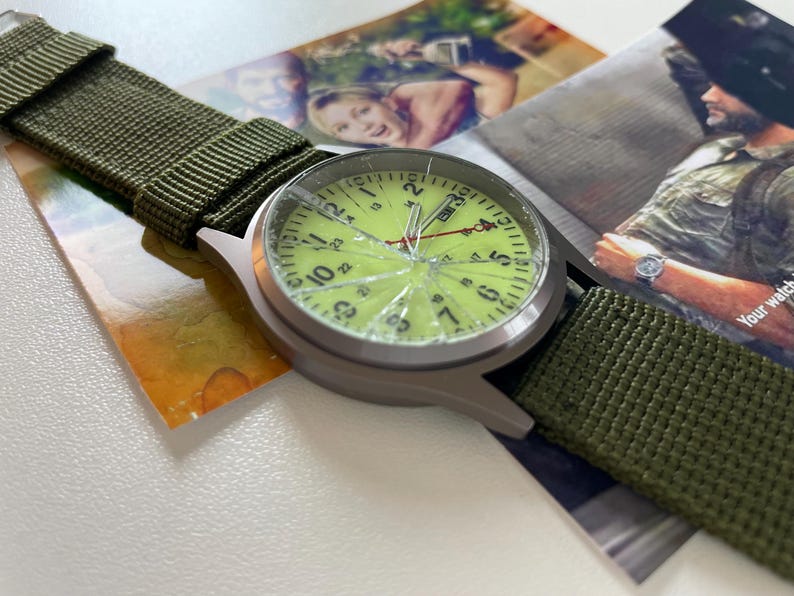 May include: A silver-toned wristwatch with a cracked, light green face and black numerals. The watch has a green nylon strap. The watch is resting on a photograph.