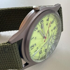 May include: A close-up of a wristwatch with a green canvas band. The watch face is a bright yellow with black numerals and a red second hand. The watch face is cracked. The watch case is a dark gray color.