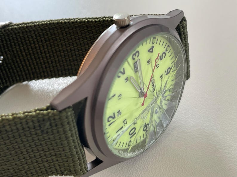 May include: A close-up of a wristwatch with a green canvas band. The watch face is a bright yellow with black numerals and a red second hand. The watch face is cracked. The watch case is a dark gray color.