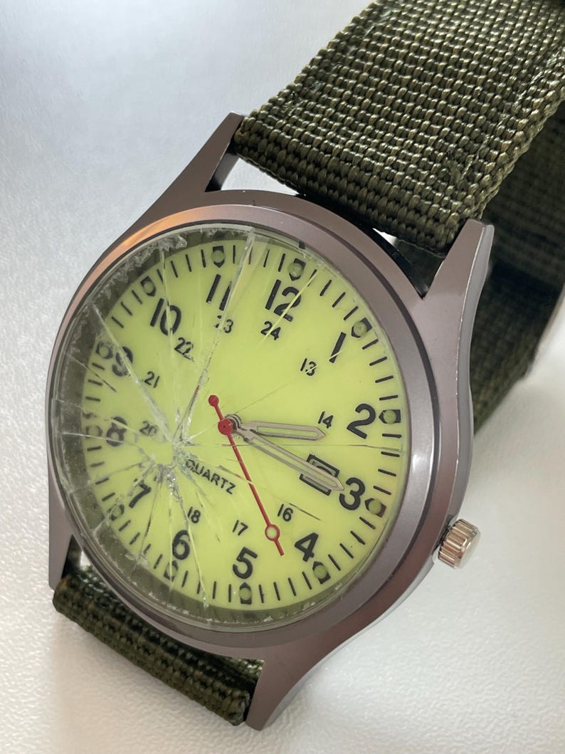 May include: A military-style wristwatch with a cracked, yellow-green face and a dark grey metal case. The watch has black numerals and hands, with a red second hand. It features a green nylon strap.
