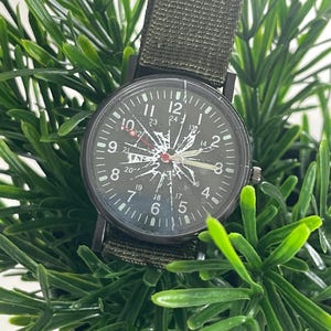 May include: A black analog wristwatch with a cracked glass face, featuring a dark dial with white numerals from 1 to 12 and smaller 24-hour markings. The watch has a textured olive green fabric strap and a black metal casing with a side crown.