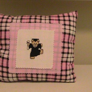 May include: Pink and black plaid pillow with a white square patch featuring a small embroidered image of a teddy bear wearing a graduation cap and gown.