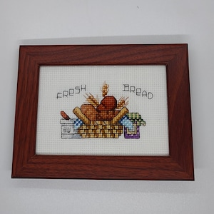 May include: A framed cross-stitch embroidery with a basket of bread, butter, and wheat. The text "FRESH BREAD" is stitched above the basket.