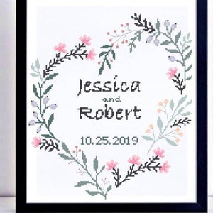 Może przedstawiać: Oprawiona haftem krzyżykowym praca z imionami "Jessica i Robert" oraz datą "10.25.2019" wewnątrz kwiatowego wieńca w kształcie serca. Wieniec zawiera r&oacute;żowe kwiaty, zielone liście i małe jagody, wszystko na białym tle.