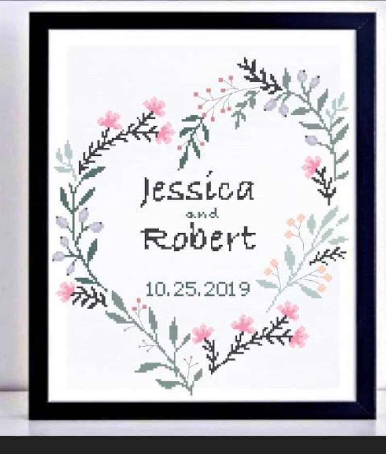 Może przedstawiać: Oprawiona haftem krzyżykowym praca z imionami "Jessica i Robert" oraz datą "10.25.2019" wewnątrz kwiatowego wieńca w kształcie serca. Wieniec zawiera r&oacute;żowe kwiaty, zielone liście i małe jagody, wszystko na białym tle.