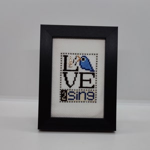 May include: A framed cross-stitch design with a blue bird singing a musical note. The design reads "LOVE SING".