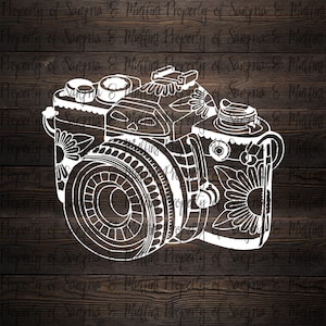 May include: A white line art decal of a camera featuring intricate floral patterns on the body and lens. The design includes detailed petals and geometric shapes etched into the camera frame, presented as a graphic illustration on a dark wood background.