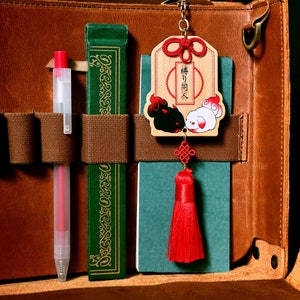 Puede incluir: Una bolsa de cuero marrón con un libro verde, un bolígrafo rojo y un amuleto de madera con un borla roja colgando. El amuleto tiene un diseño de animal blanco y negro y el texto "神 々" en japonés.