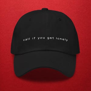 May include: Black baseball cap with the text "call if you get lonely" in white. The cap has a curved brim and a button on top. The background is a solid red.