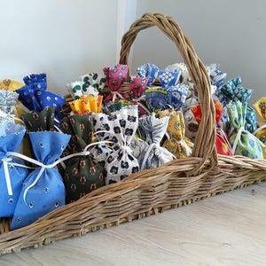 Może przedstawiać: Koszyk wiklinowy wypełniony małymi woreczkami z tkaniny w różnych kolorach i wzorach. Woreczki są związane białą wstążką i mają falbankę.