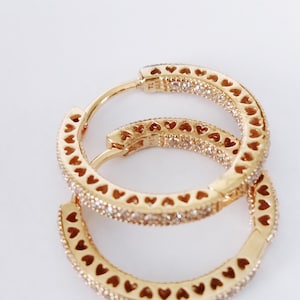 May include: A pair of gold hoop earrings with a heart design and small clear stones. The earrings are made of metal and have a polished finish.