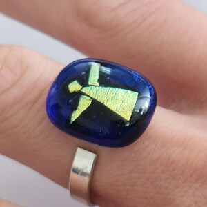 May include: A ring featuring a smooth, rectangular, deep blue glass cabochon with rounded edges. Embedded within the glass is a shimmering, iridescent yellow-green figure that resembles an angel with outstretched wings and a triangular body.