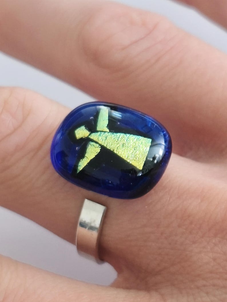 May include: A ring featuring a smooth, rectangular, deep blue glass cabochon with rounded edges. Embedded within the glass is a shimmering, iridescent yellow-green figure that resembles an angel with outstretched wings and a triangular body.