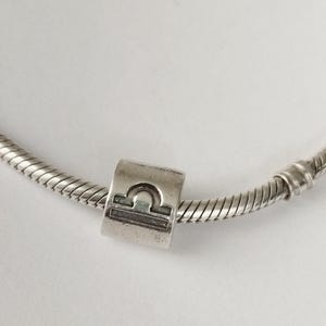 Può includere: Un charm d'argento con il simbolo zodiacale della Bilancia, una bilancia stilizzata. Il charm cilindrico è posto su una catena a serpente d'argento. Lo sfondo è una superficie bianca.