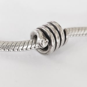 Può includere: Un ciondolo a forma di serpente color argento avvolto attorno a un bracciale a catena flessibile. Il ciondolo presenta una testa di serpente dettagliata con piccoli occhi e un corpo testurizzato che si avvolge attorno alla catena in tre anelli.