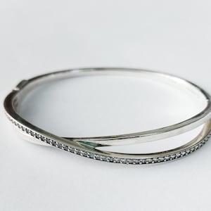 Peut inclure: Un bracelet jonc argenté avec un design torsadé. Une partie est ornée de petits pierres précieuses transparentes. Le bracelet est présenté sur un fond blanc, soulignant son élégance.