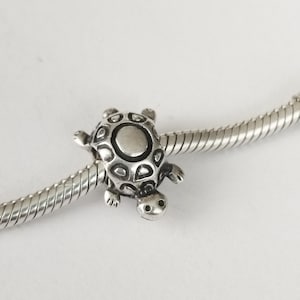 Può includere: Un charm a forma di tartaruga argentata con guscio e arti dettagliati. Il charm è attaccato a una catena d'argento, creando un tema nautico. La tartaruga ha un motivo a guscio dettagliato e un volto sorridente. Questo charm è adatto per bracciali e collane.