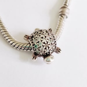 Può includere: Un charm a forma di tartaruga argentata con un guscio di perline e piccoli accenti turchesi. Il charm è attaccato a un braccialetto a catena a serpente argentato, creando un tema nautico. Il guscio della tartaruga è incrostato di pietre chiare e turchesi.