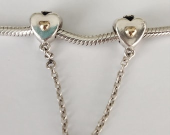 May include: Silver-toned charm with two heart-shaped pendants, each featuring a gold-colored heart in the center. The pendants are connected by a delicate chain, designed to be attached to a bracelet or necklace.
