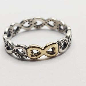 Op de afbeelding: Een zilverkleurige ring met een oneindigheidssymboolontwerp. De ring heeft een goudkleurig oneindigheidssymbool in het midden, de rest van de band is gemaakt van zilverkleurig metaal. De ring staat op een witte achtergrond.