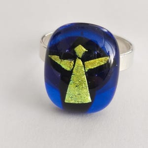 May include: A ring featuring a smooth, oval-shaped glass cabochon in deep blue with a central, stylized yellow-green figure resembling an angel with outstretched arms. The glass piece is mounted on a simple, polished silver-toned metal band.
