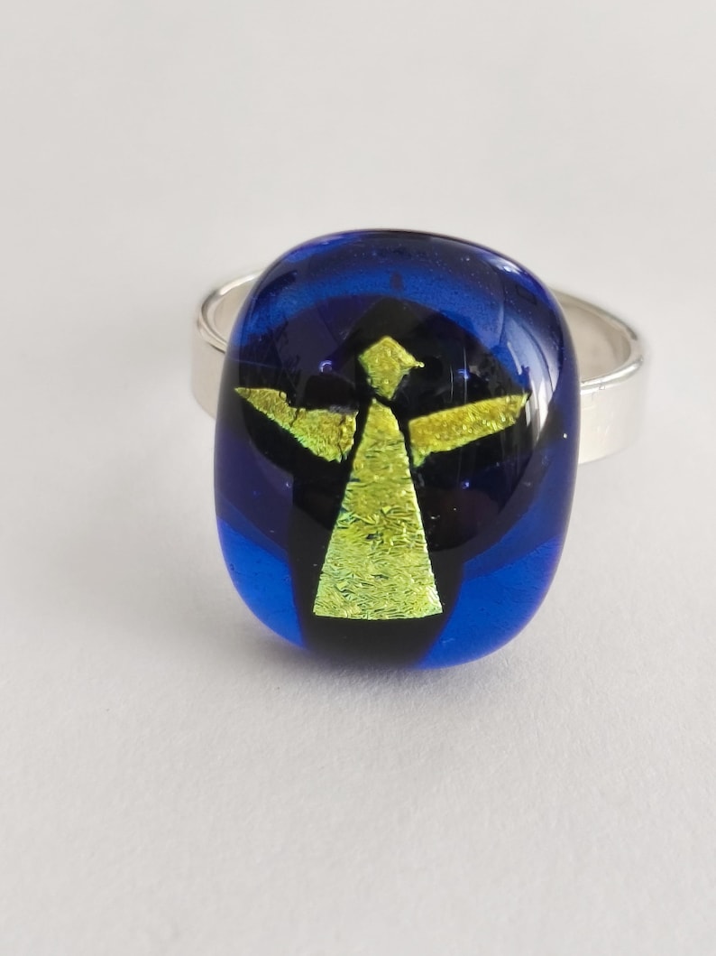 May include: A ring featuring a smooth, oval-shaped glass cabochon in deep blue with a central, stylized yellow-green figure resembling an angel with outstretched arms. The glass piece is mounted on a simple, polished silver-toned metal band.