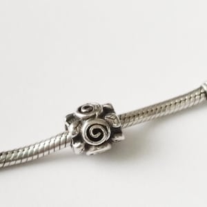 Può includere: Una perlina charm in argento con un design a rosa dettagliato. La perlina è di forma quadrata con una rosa al centro e foglie intorno. Il charm è infilato su una catena a serpente d'argento, su sfondo bianco.