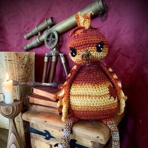 May include: A crocheted bird toy with orange, red and yellow stripes. The bird has a crown on its head and is sitting on a wooden chest with black metal accents.