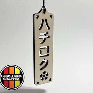 May include: A wooden sign with the Japanese characters "ハチクロ" carved into it. The sign is hanging from a black string and has a small flower design at the bottom.