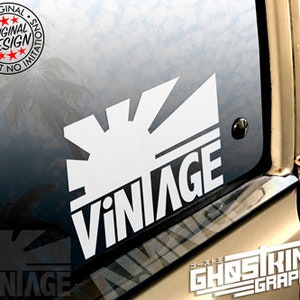 May include: White vinyl sticker with a stylized sun and the word "Vintage" in bold, white letters. The sticker is on a car window.