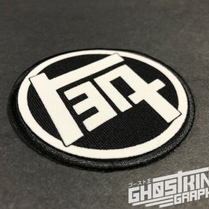 May include: A black and white embroidered patch with a stylized "T" logo. The patch is circular and has a white border.