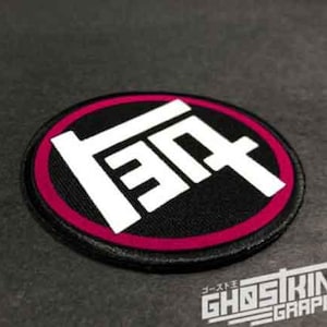 May include: A circular embroidered patch featuring a black background with a magenta border and a large white stylized Japanese character logo in the center. The patch has a stitched edge and a flat, textured surface design.
