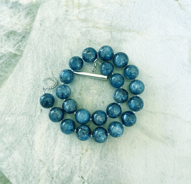 K&ouml;nnte beinhalten: Eine Halskette aus blauen Edelsteinperlen mit einem silbernen Verschluss. Die Perlen sind rund und haben eine leicht strukturierte Oberfl&auml;che.