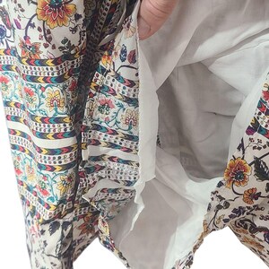 May include: A white dress with a floral pattern in yellow, orange, and purple. The dress has a white lining and a decorative border with a repeating pattern. The fabric is lightweight and appears to be suitable for warm weather.