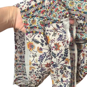 May include: Cream-colored fabric with a floral pattern in yellow, red, and blue. The fabric has a bohemian style with a mix of floral and geometric designs. The garment appears to be a top or dress.