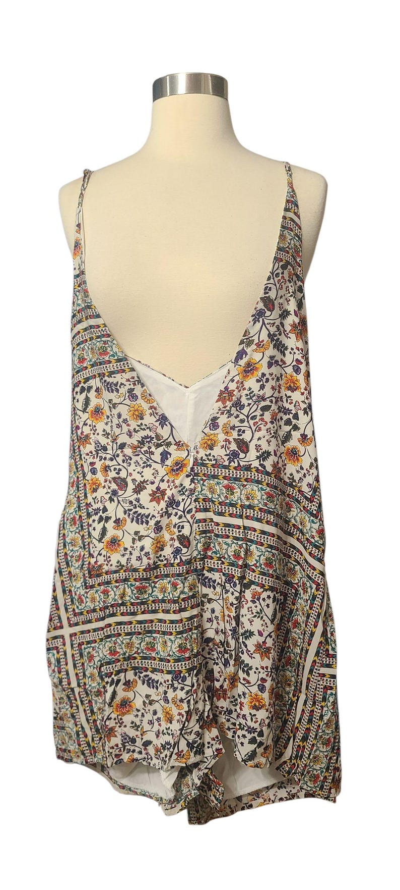 May include: A floral print romper with a deep V-neck and spaghetti straps. The romper features a white background with a colorful floral pattern in yellow, red, and blue. The garment is displayed on a mannequin.