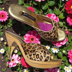 May include: Two Betsy Johnson platform mules with a gold metallic finish and leopard print accents. The open-toe heels have a wooden platform and a gold stiletto heel. The shoes are set against a backdrop of pink flowers and greenery.