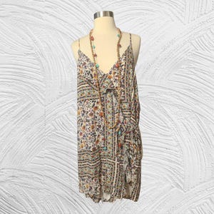 May include: A floral print dress with spaghetti straps and a V-neck. The dress features a cream-colored background with a pattern of flowers in shades of orange, yellow, and blue. A long beaded necklace with turquoise and orange beads is draped over the dress.