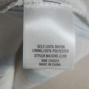 以下が含まれることがあります： 黒い文字が書かれた白い布タグ。タグには「SELF: 100% RAYON, LINING: 100% POLYESTER, STYLE# M4169D-11FR, RN# 150025, MADE IN CHINA」と書かれています。タグは白と青のストライプの生地に付いています。